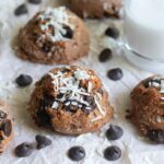 chocolate chip cookies topped with shredded coconut, arranged on parchment paper with extra chocolate chips scattered around.