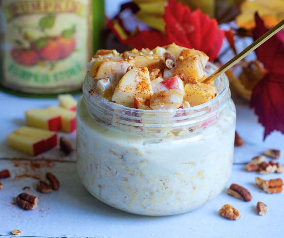 A glass jar filled with creamy overnight oats topped with fresh apple chunks, pecans, and a sprinkle of cinnamon, surrounded by autumn leaves and apple slices.