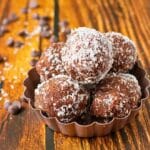 Cookie Dough Bitescoated in shredded coconut, arranged in a small brown dish on a rustic wooden surface, with scattered chocolate chips in the background.