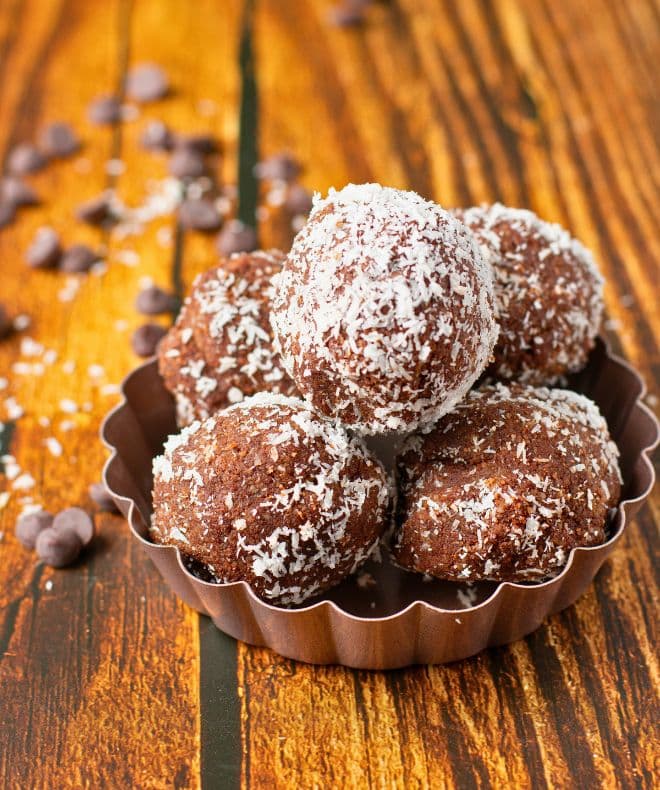 Cookie Dough Bitescoated in shredded coconut, arranged in a small brown dish on a rustic wooden surface, with scattered chocolate chips in the background.