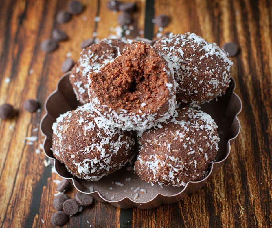 Chocolate almond balls coated in shredded coconut, arranged in a small brown dish on a rustic wooden surface, with scattered chocolate chips in the background.