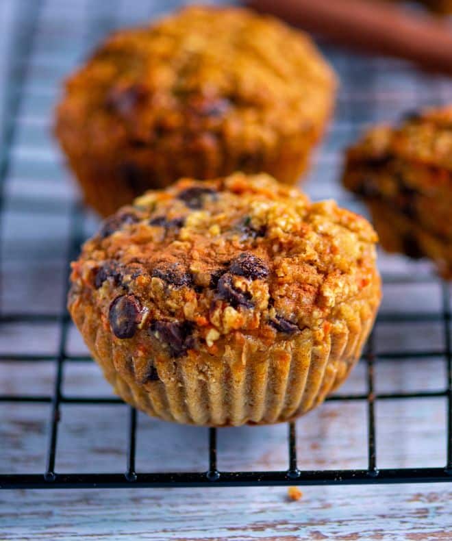 Carrot oat muffin with a golden, textured top and visible oats and chocolate chips, resting on a wire cooling rack with softly blurred muffins in the background.