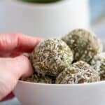 Hand reaching for homemade matcha energy bites rolled in coconut flakes in a white bowl