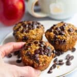 a hand holding one of 3 Gluten-Free Oatmeal Apple Muffins on a white plate with chocolate chips around and an apple on the background