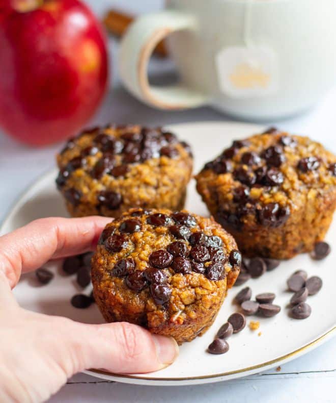 a hand holding one of 3 Gluten-Free Oatmeal Apple Muffins on a white plate with chocolate chips around and an apple on the background