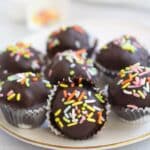 Chocolate-covered Protein Brownie Bites topped with rainbow sprinkles and served in mini paper liners on a white plate.