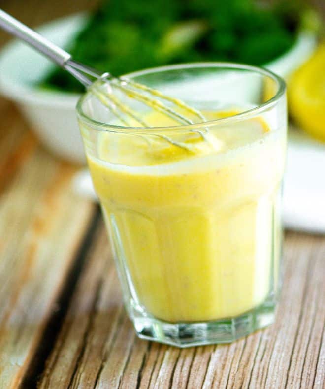 Creamy high-protein lemon egg salad dressing in a clear glass, with a whisk lifting the thick, smooth dressing. Fresh lemon halves and a bowl of leafy greens are blurred in the background on a wooden table.