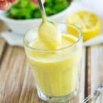 Creamy high-protein lemon egg salad dressing in a clear glass, with a spoon lifting the thick, smooth dressing. Fresh lemon halves and a bowl of leafy greens are blurred in the background on a wooden table.